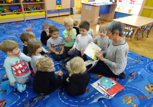 Dzieci siedzą w kole, pani bibliotekarka czyta dzieciom książkę.